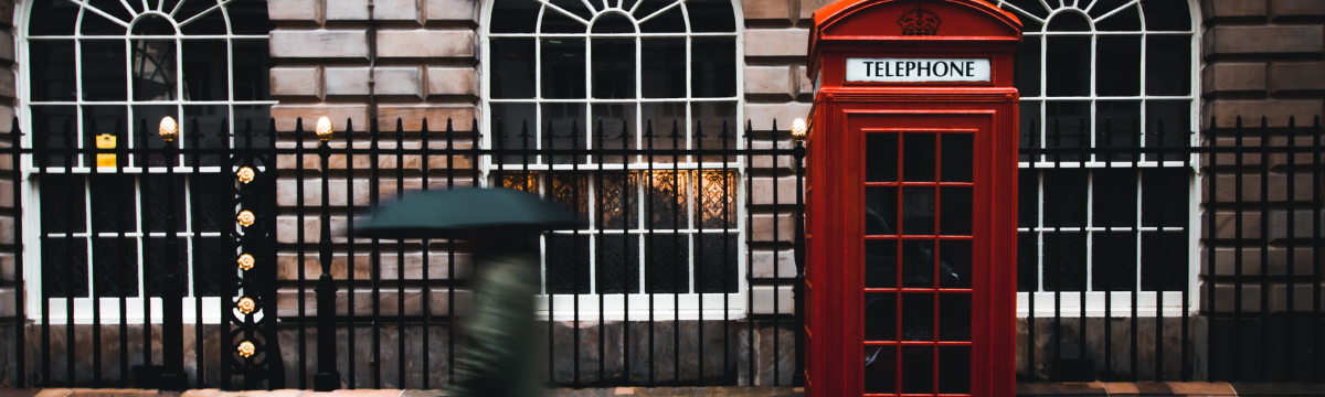 Image of a London phone box on a street. A person holding an umbrella is walking by.