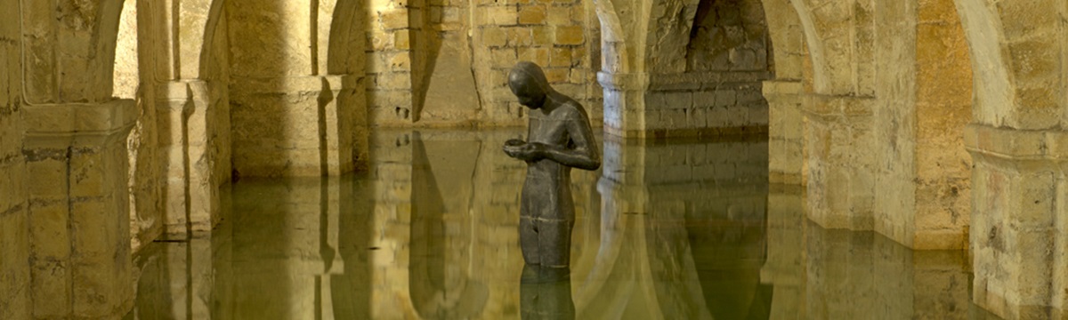Jumbotron Image - a view of the Cathedral crypt at Winchester Cathedral. It is partially flooded, and a statue of a man - Antony Gormley Sound II - stands, contemplating the water in its cupped hands.