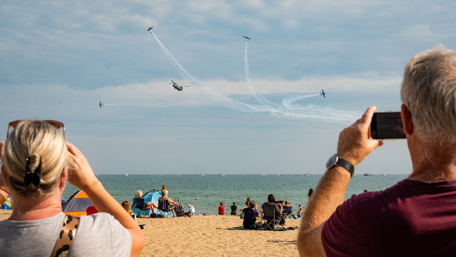 2 people on the beach video recording planes in the air at Bournemouth Air Festival; other groups of people are sitting on the beach watching the show.