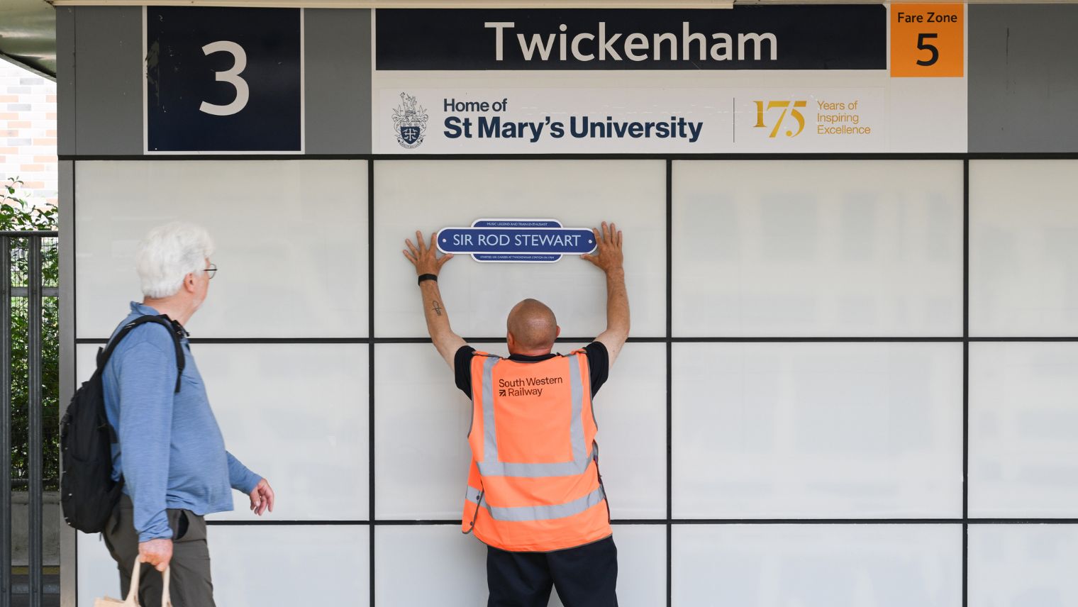 Man in Hi-vis tabard placing a plaque on a grey wall while a passer-by looks on