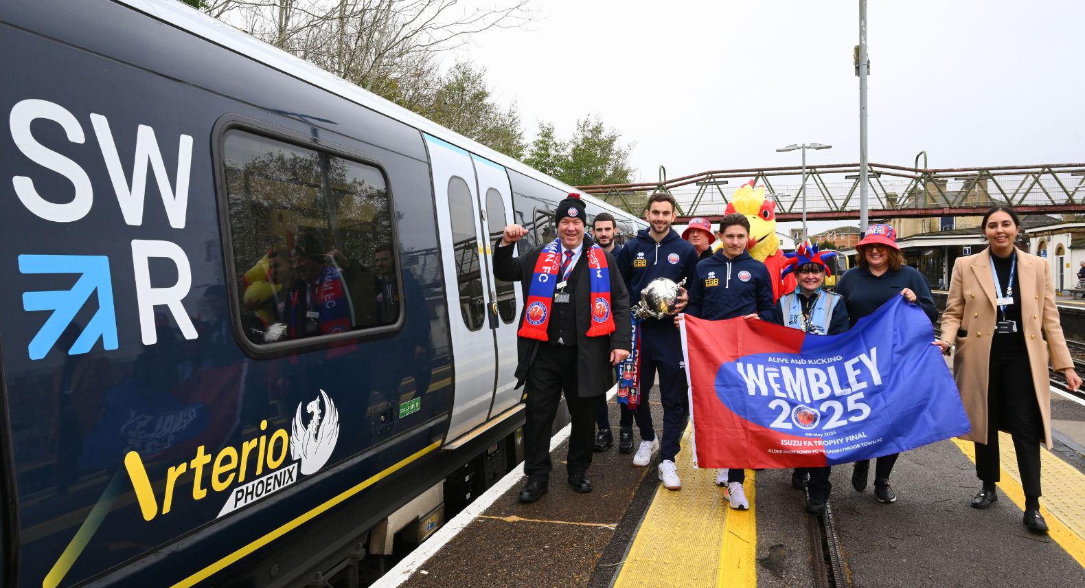 Group of people holding a banner saying Wembley 2025 walking beside a blue train with SWR Arterio Phoenix on the side