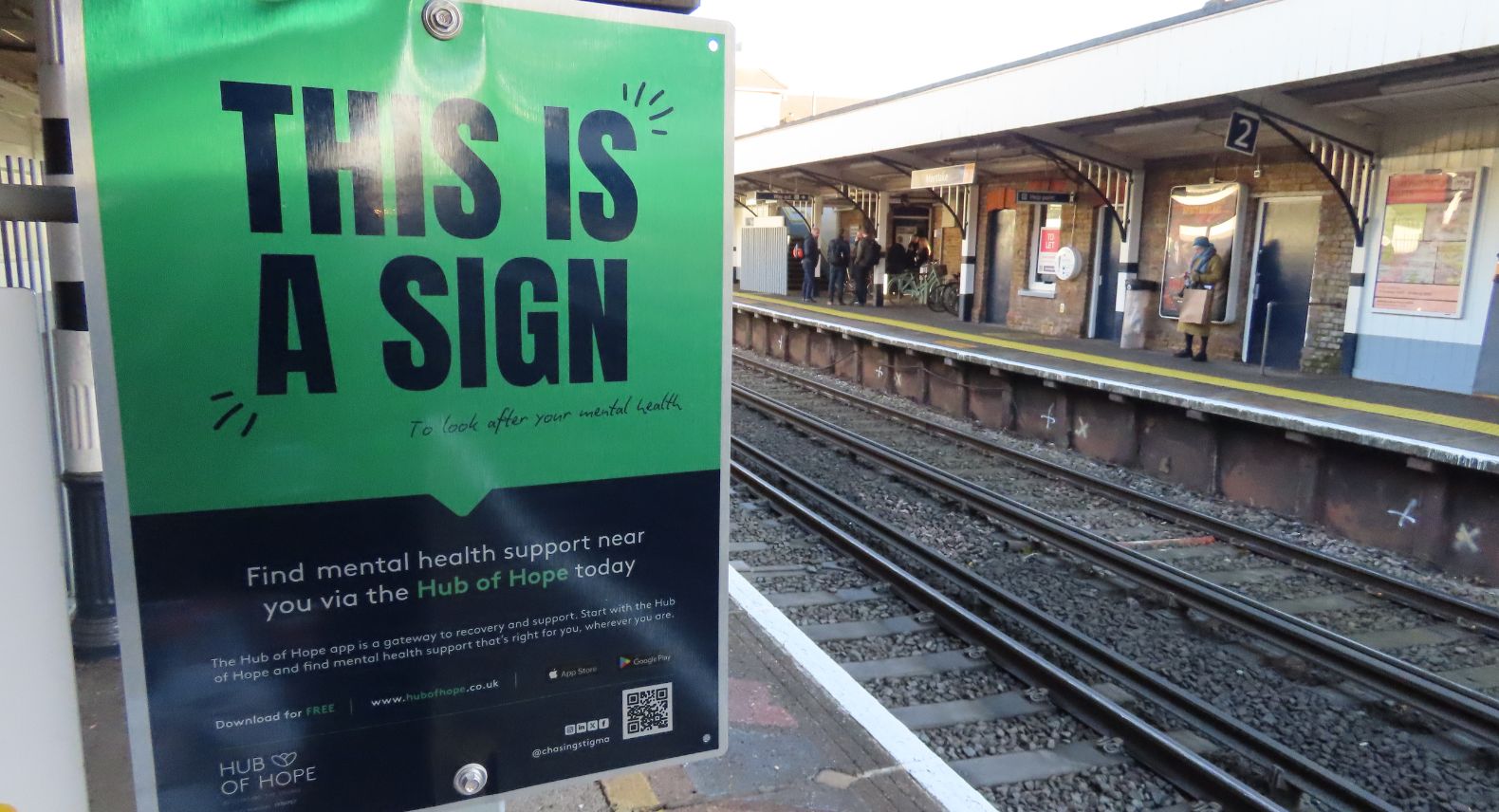 A large green and black poster placed on a station platform saying in Block capitals 'This is a sign' with the rail tracks and another platform in the background