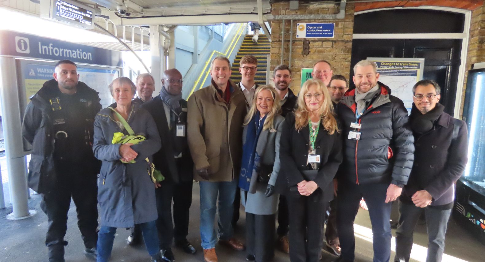 A group of people standing on a station platform in from of a set of stairs and a sign saying 'Information'