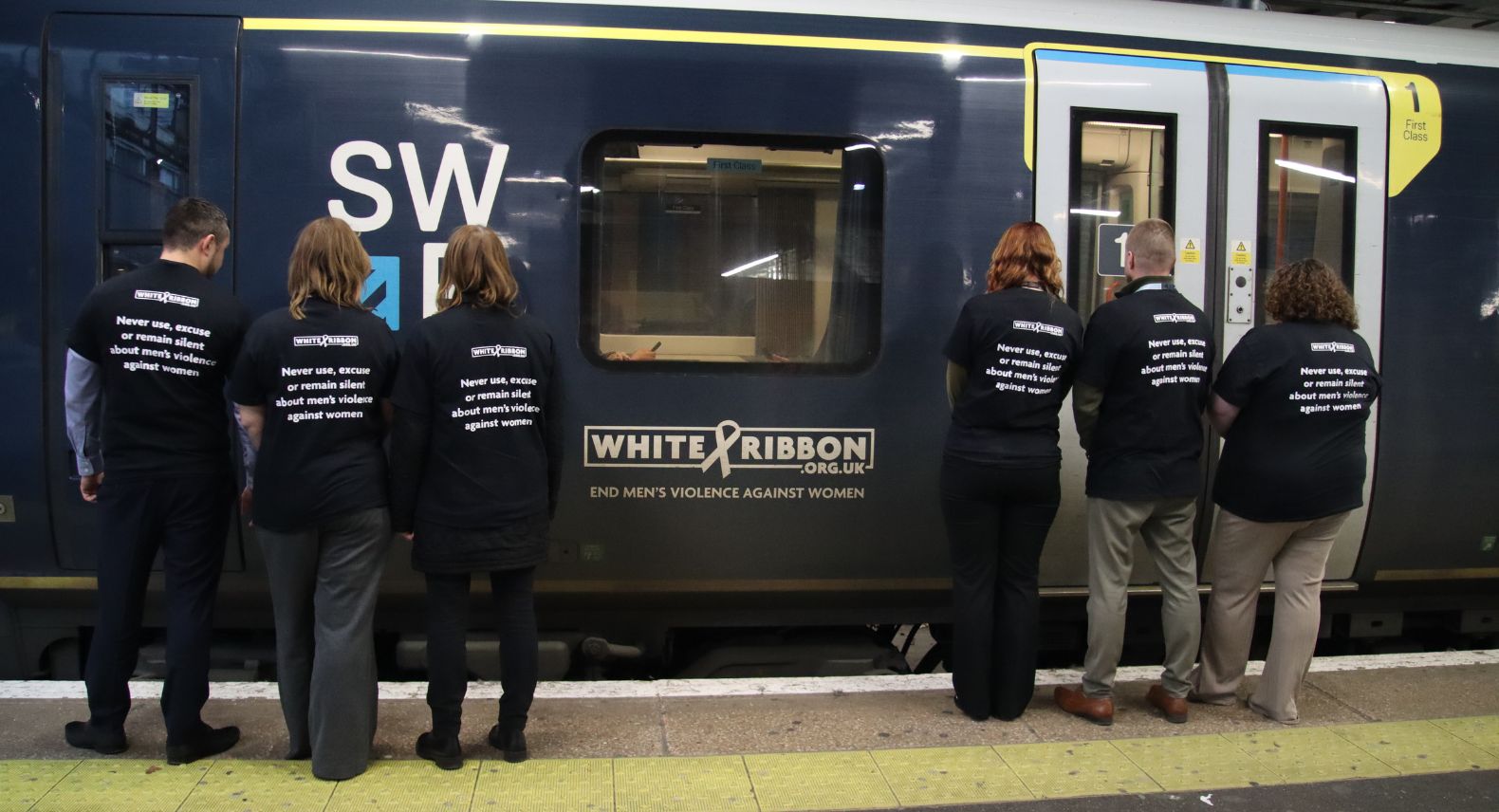 A group of six people with backs to the camera wearing t-shirts which say "Never use, excuse or remain silent about men's violence against women"
