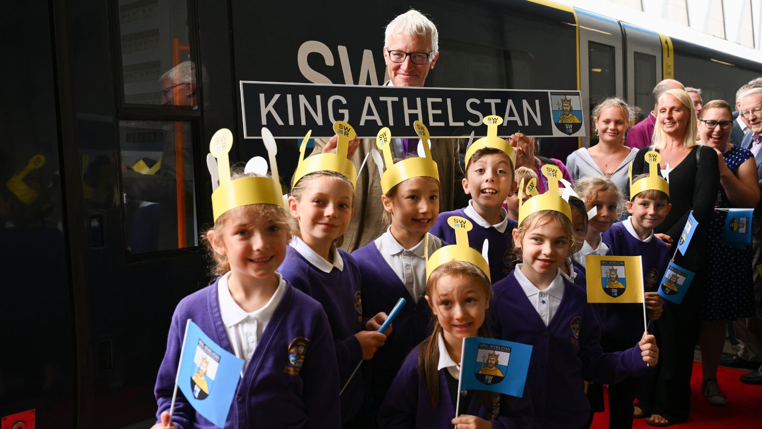 Group of school children wearing crowns and waving flags standing in front of a train