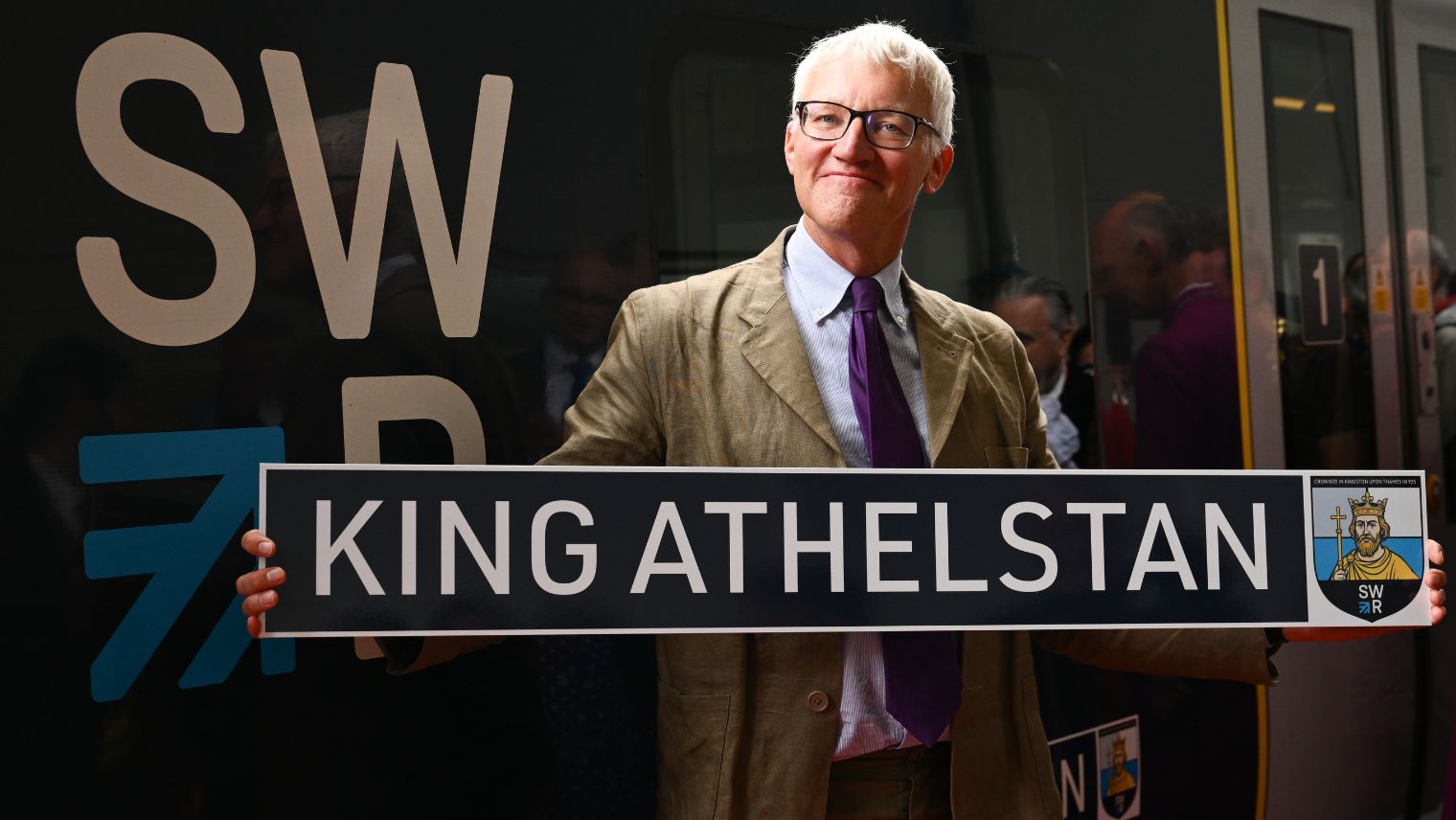 Grey haired man in glasses standing in front of a train with an SWR logo, holding a name plate saying King Athelstan