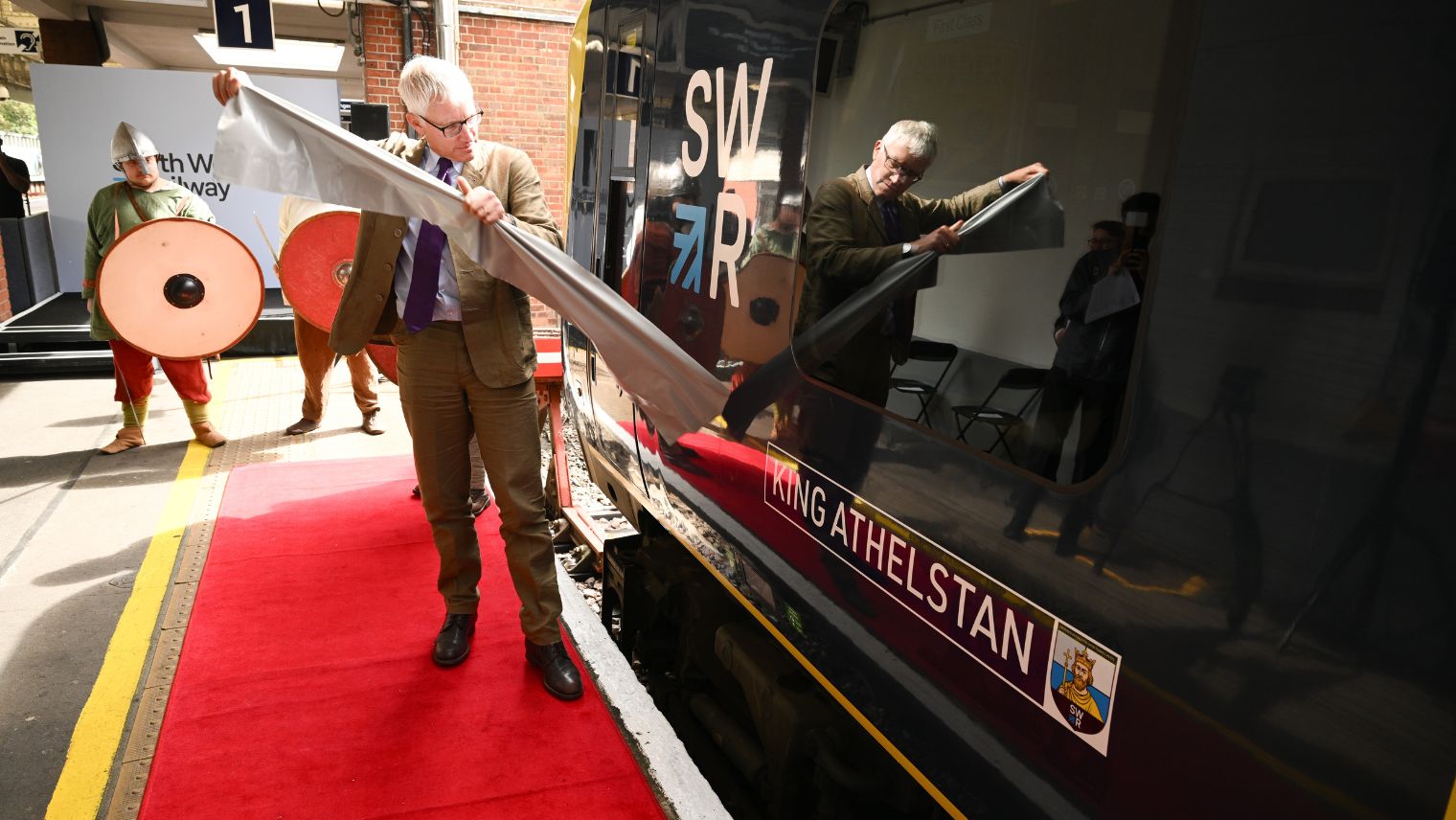 Grey haired man in glasses standing in front of a train with an SWR logo, pulling off a sticker to reveal a name plate saying King Athelstan