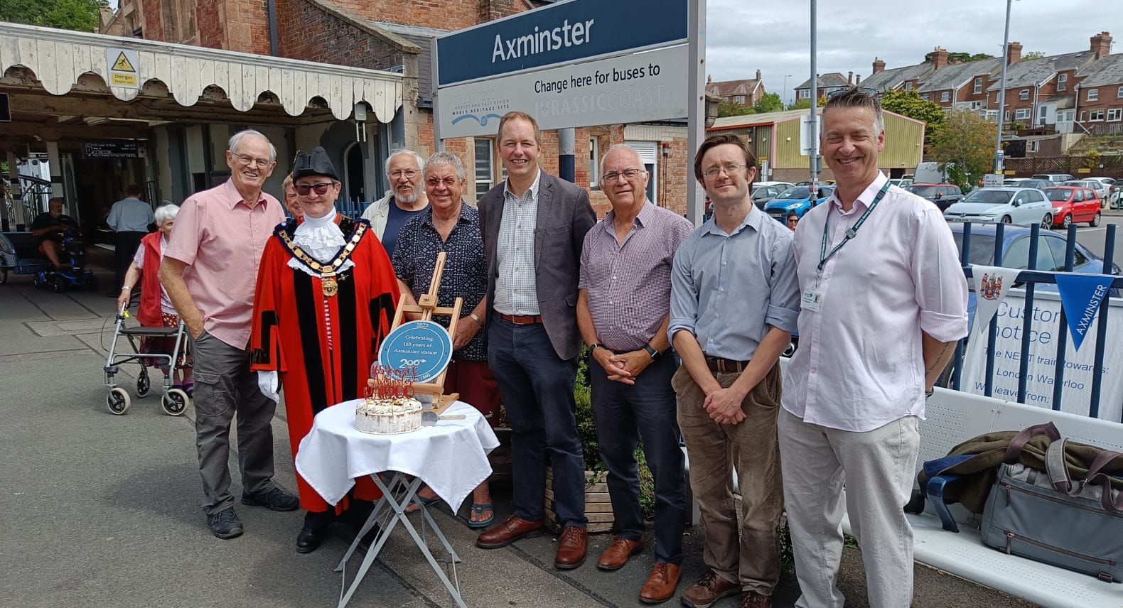 Group of 8 people (including one in Mayoral robes) standing behind a small table with a round blue plaque and a cake on it in front of a platform sign saying Axminster