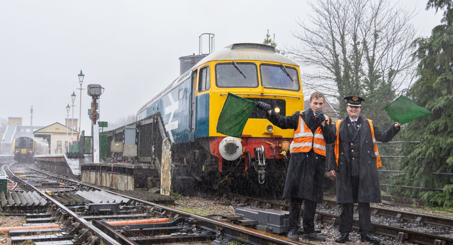 Two people in railway uniforms and hi-vis waving green flags standing in front of a locomotive