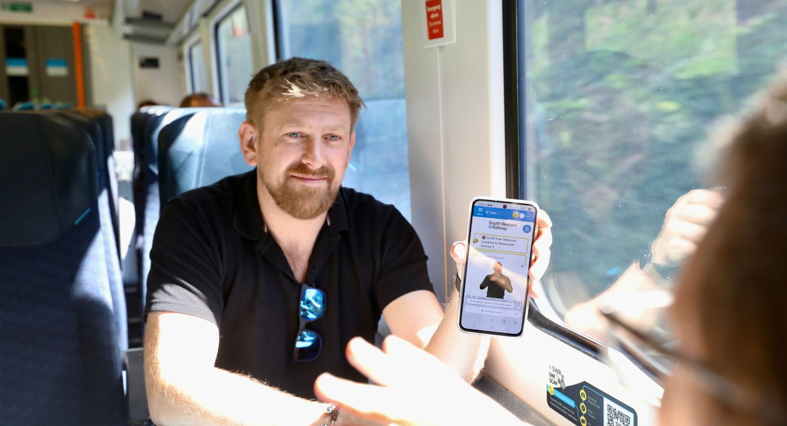 Man sitting on train showing mobile phone to someone opposite