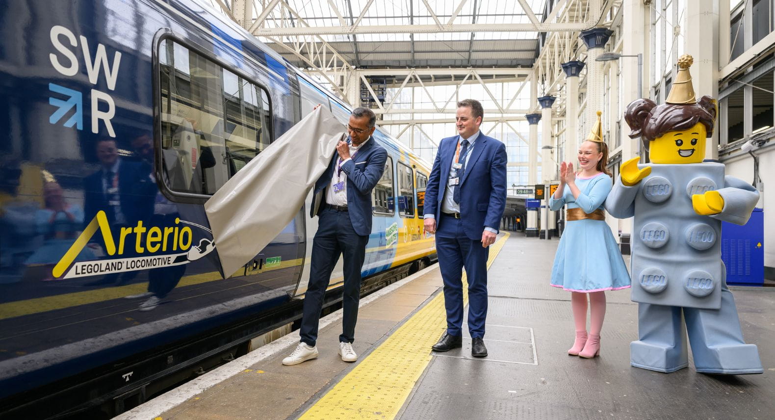 Two people remove a sticker from the side of an Arterio train to reveal 'The LEGOLAND Locomotive' while a girl and adult-size LEGO mini figure look on
