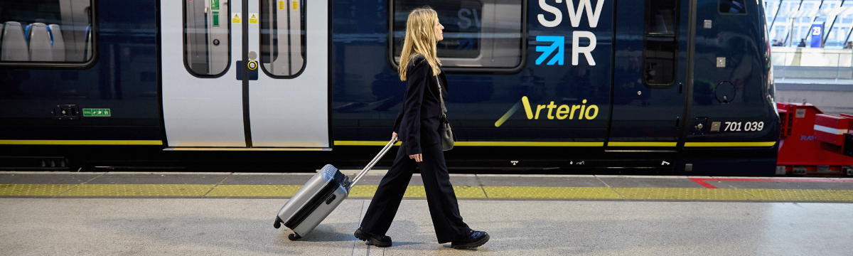 A woman dressed in dark clothing walks along a train platform, pulling a silver suitcase past a stationary South Western Railway Arterio train with “SWR” branding and blue and yellow detailing.