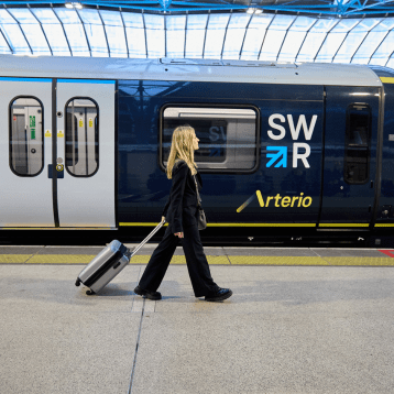 A woman dressed in dark clothing walks along a train platform, pulling a silver suitcase past a stationary South Western Railway Arterio train with “SWR” branding and blue and yellow detailing.