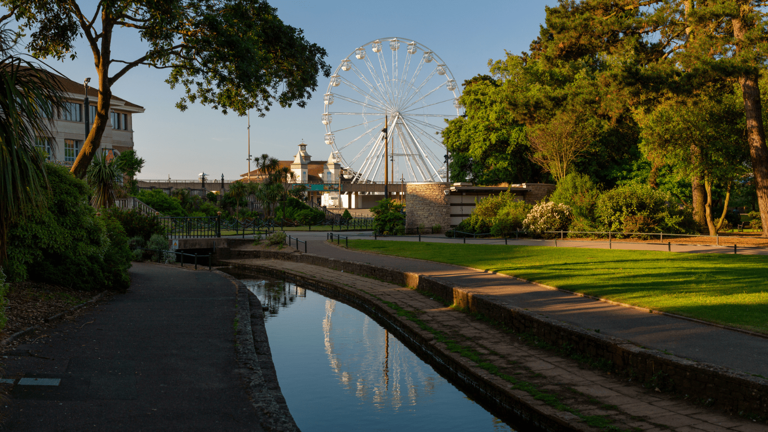 A canal-side path lined with trees, with Bournemouth’s Ferris wheel rising behind buildings and reflecting in the water.