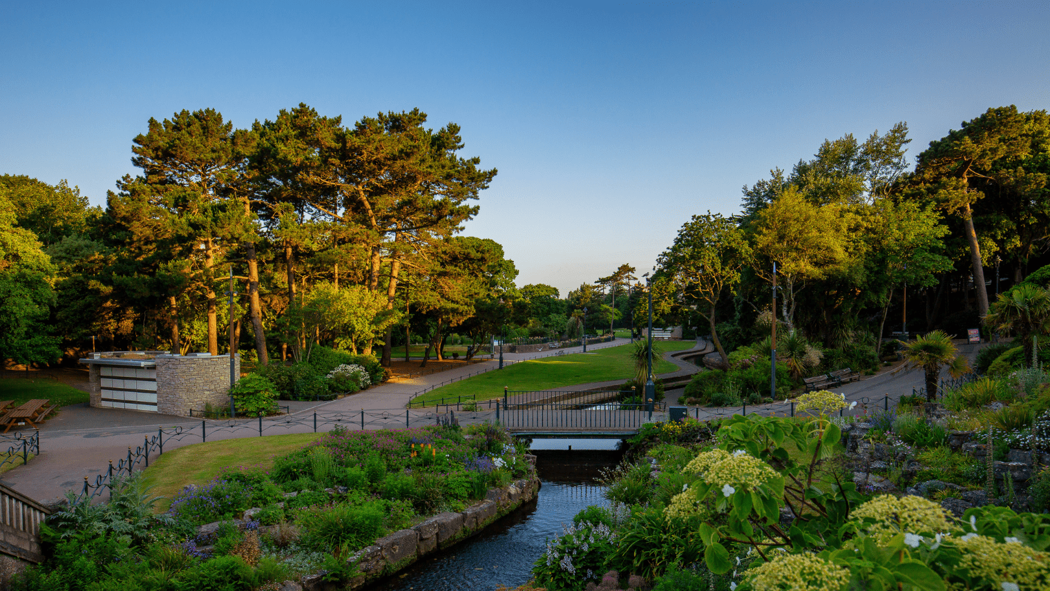 A landscaped urban park on a sunny day, featuring a gently curving stream crossed by a metal pedestrian bridge. Flowering plants and lush greenery fill garden beds on either side, bordered by paved walkways with benches and lampposts. Tall trees provide shade, and a small stone building sits to the left, surrounded by vibrant foliage.