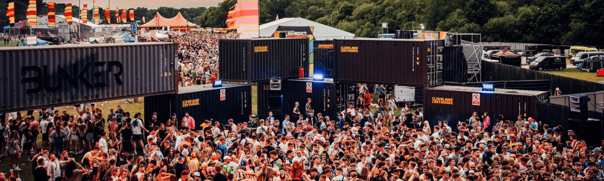 Crowd dancing in a forest under colorful geometric canopy at a psychedelic DJ stage.