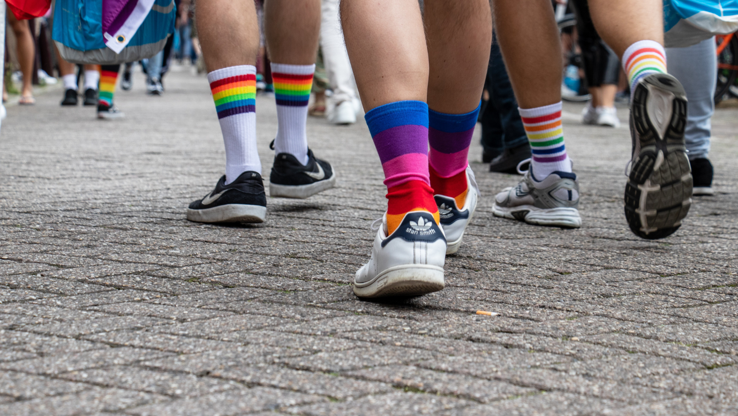 Image of 3 people's feet and calves. They're wearing colourful socks and walking on the street at a Pride event