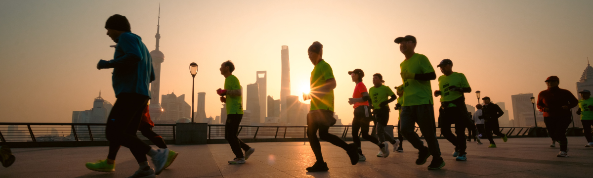 Runners silhouetted against a sunrise skyline, jogging along a waterfront promenade with long shadows.