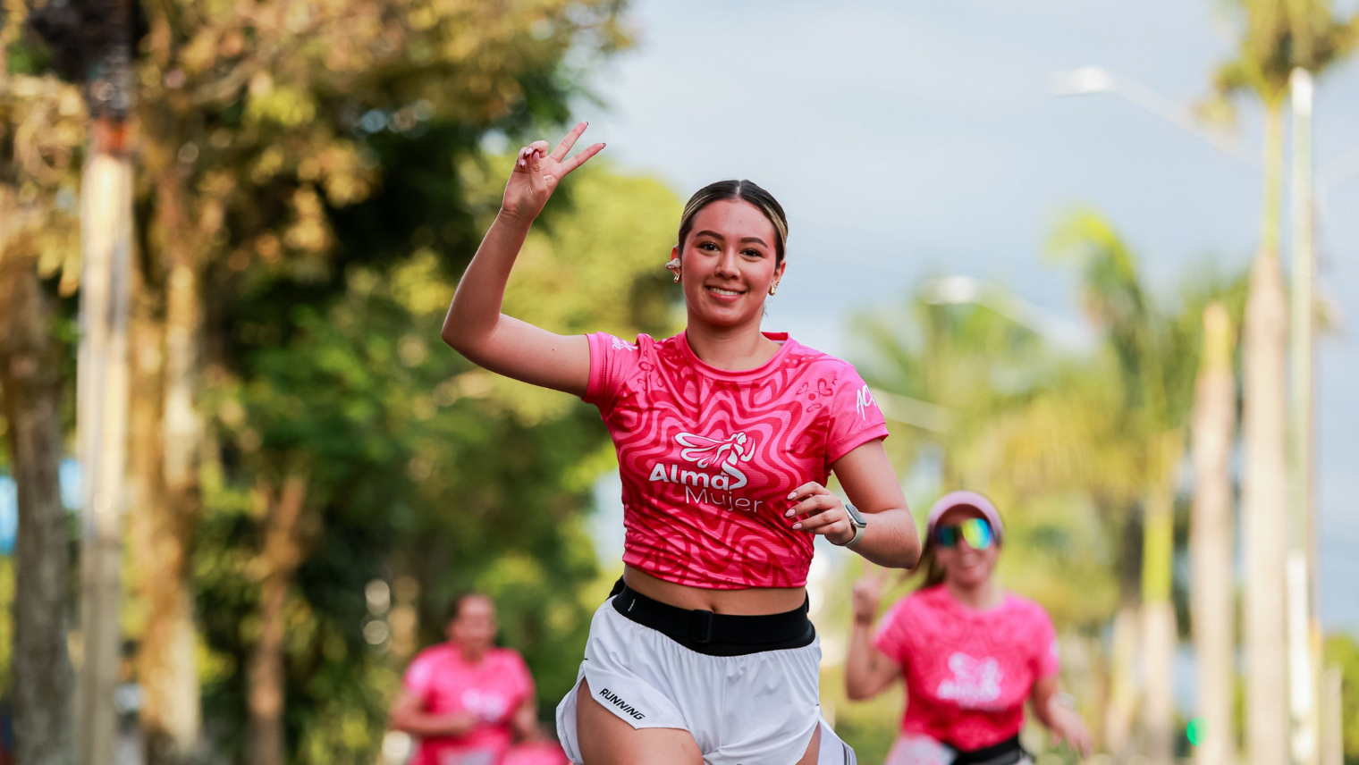 Woman in bright pink “Alma Mujer” top and white shorts flashing a peace sign while running on a sunny, palm‑lined street.