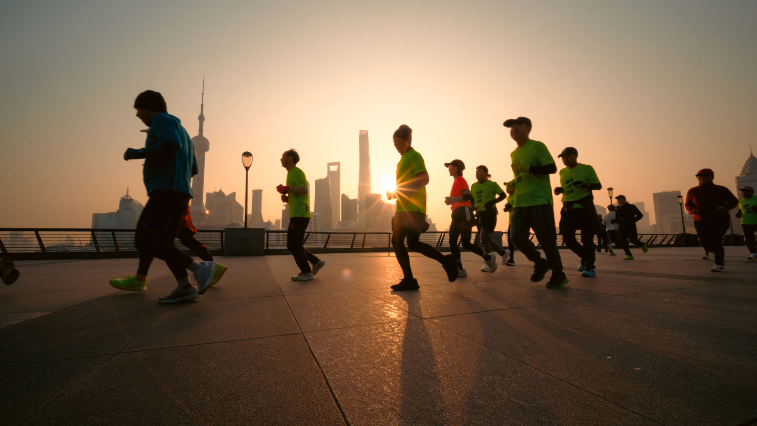 Runners silhouetted against a sunrise skyline, jogging along a waterfront promenade with long shadows.