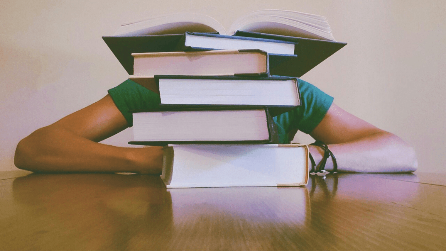 A person sits at a table with a tall stack of books hiding their face.
