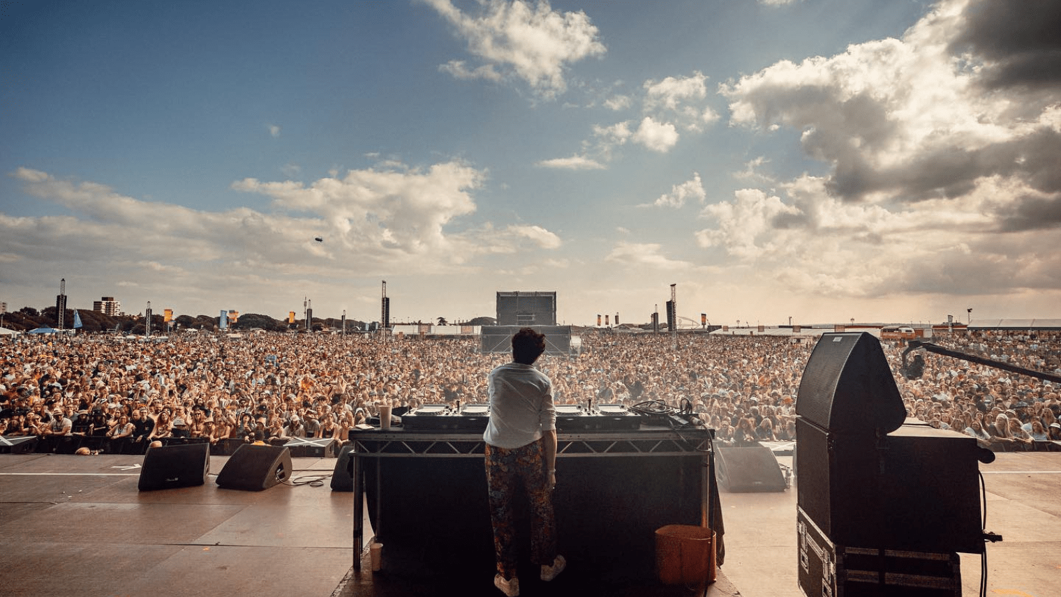 DJ performing to a huge crowd at a seaside festival, with sunlight breaking through clouds over the stage.