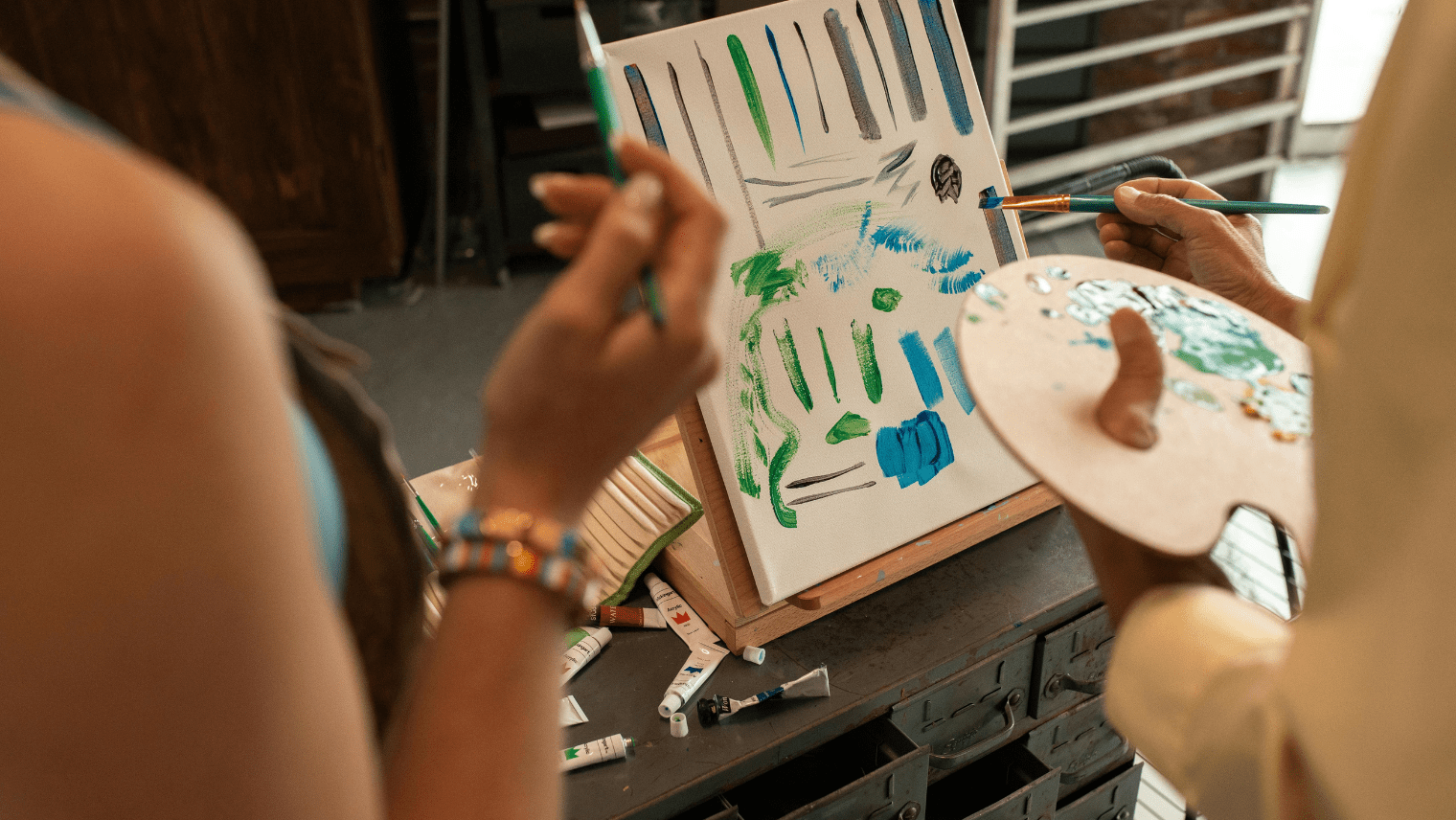 Two people paint on a canvas covered with colorful brushstrokes in green, blue, and purple; paint tubes and a palette rest nearby.