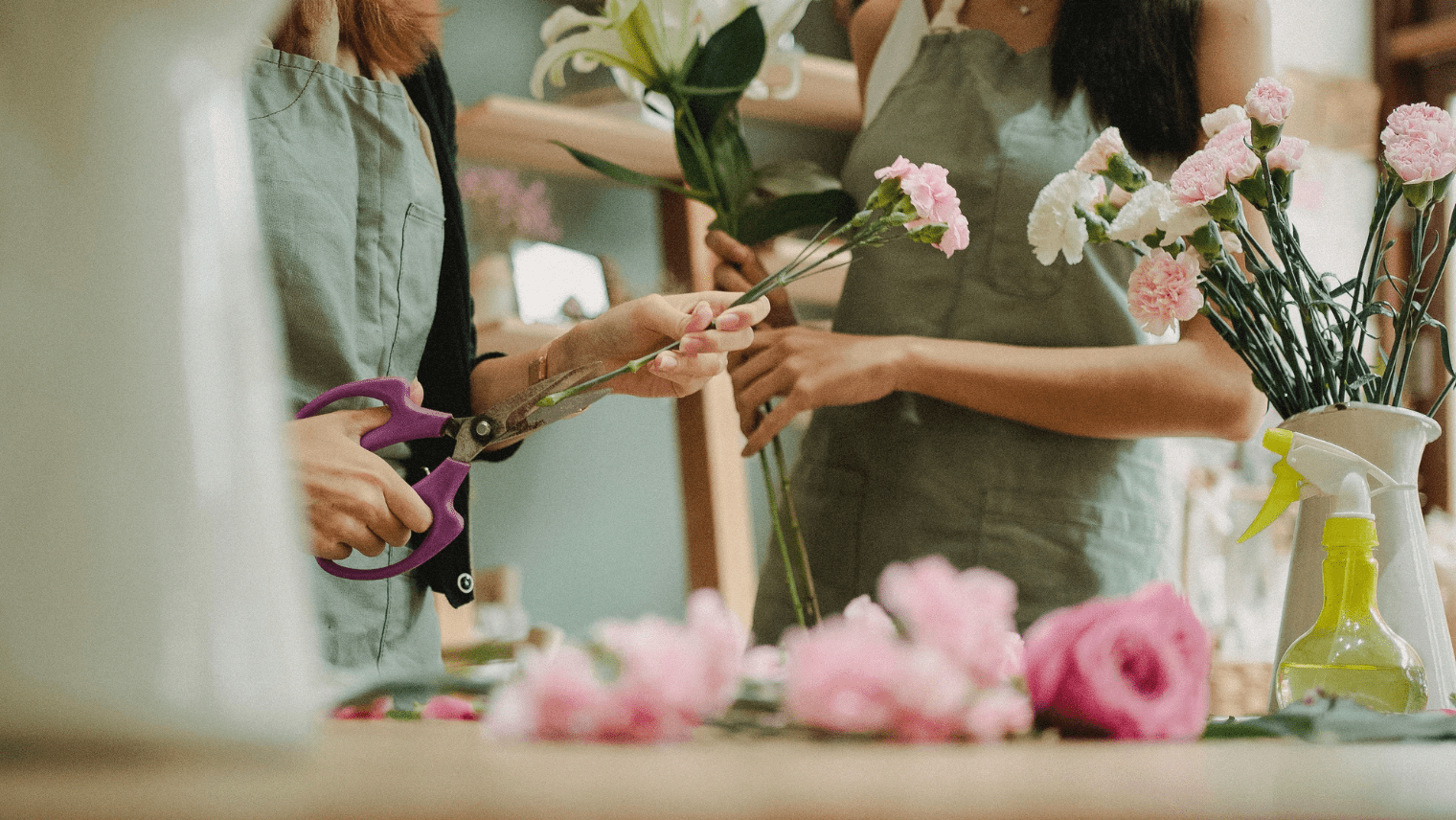 Two people wearing light green aprons arrange flowers; one cuts pink carnations with purple scissors while the other holds white lilies and carnations. Pink roses lie on the table in front of them.
