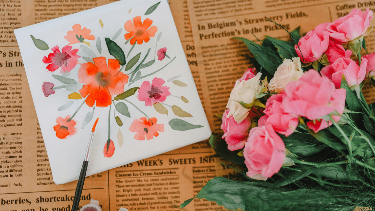 A floral watercolor painting with red, orange, and pink flowers rests on a newspaper beside a bouquet of fresh pink roses and a paintbrush with an orange tip.