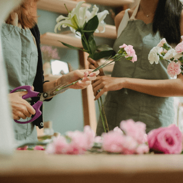 Two people wearing light green aprons arrange flowers; one cuts pink carnations with purple scissors while the other holds white lilies and carnations. Pink roses lie on the table in front of them.