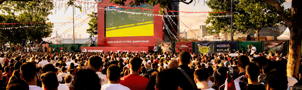 Crowd watching a football match on a big outdoor screen at a fan zone.
