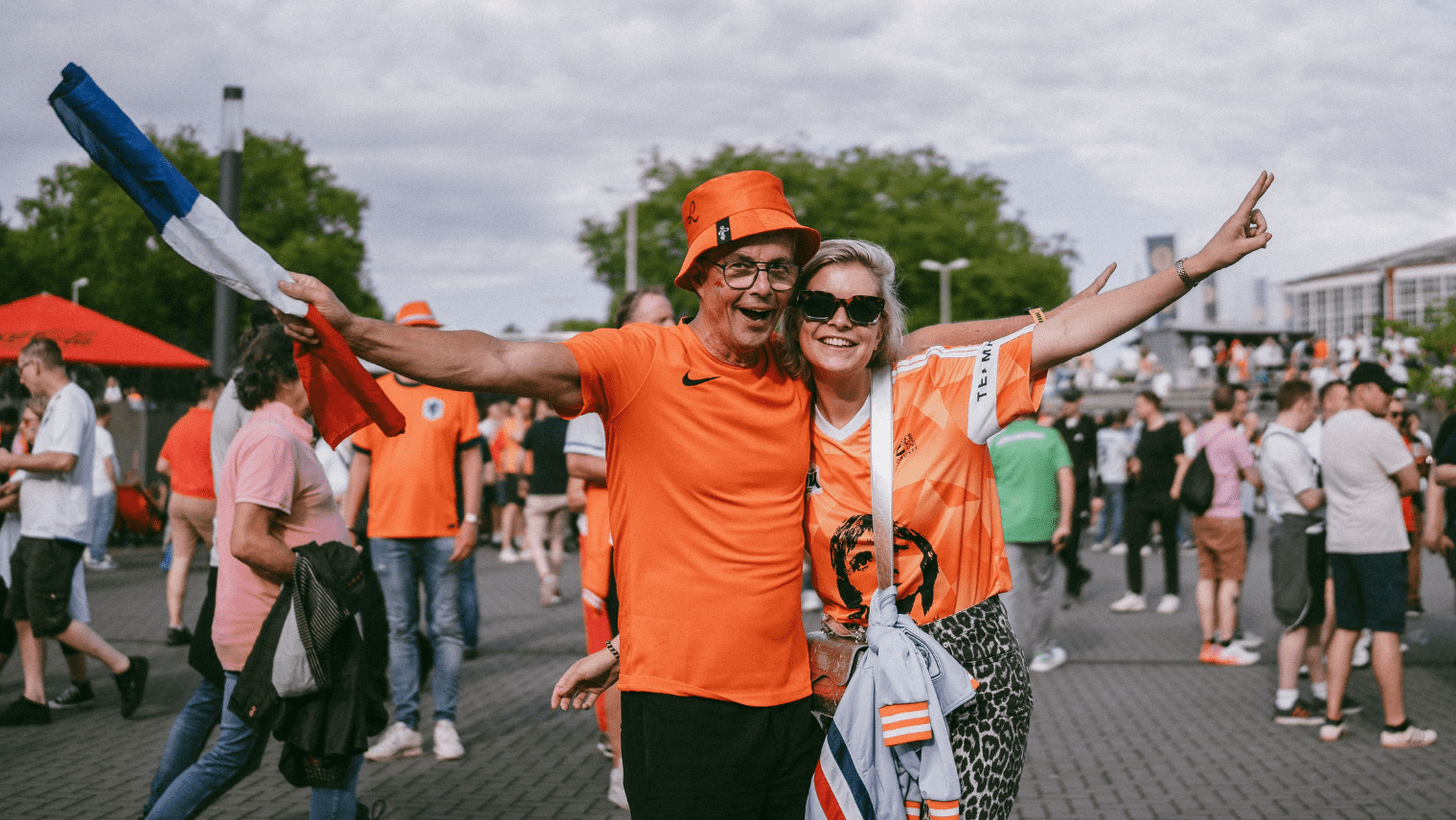 Two fans in orange outfits celebrating outdoors with a Netherlands flag among a crowd.