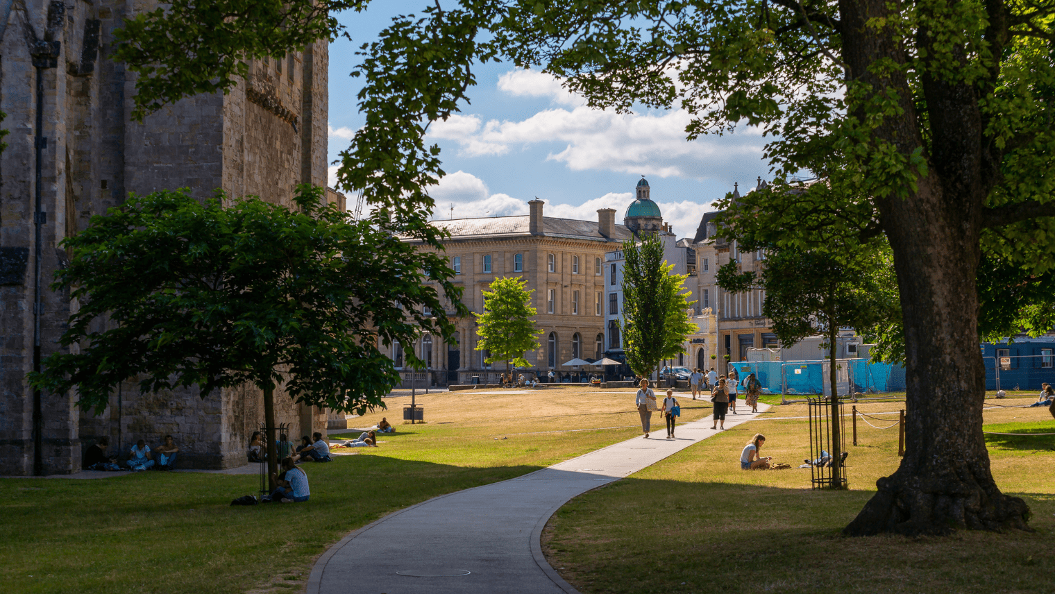 A curved path runs through a sunny park where people sit and walk among historic buildings and trees.