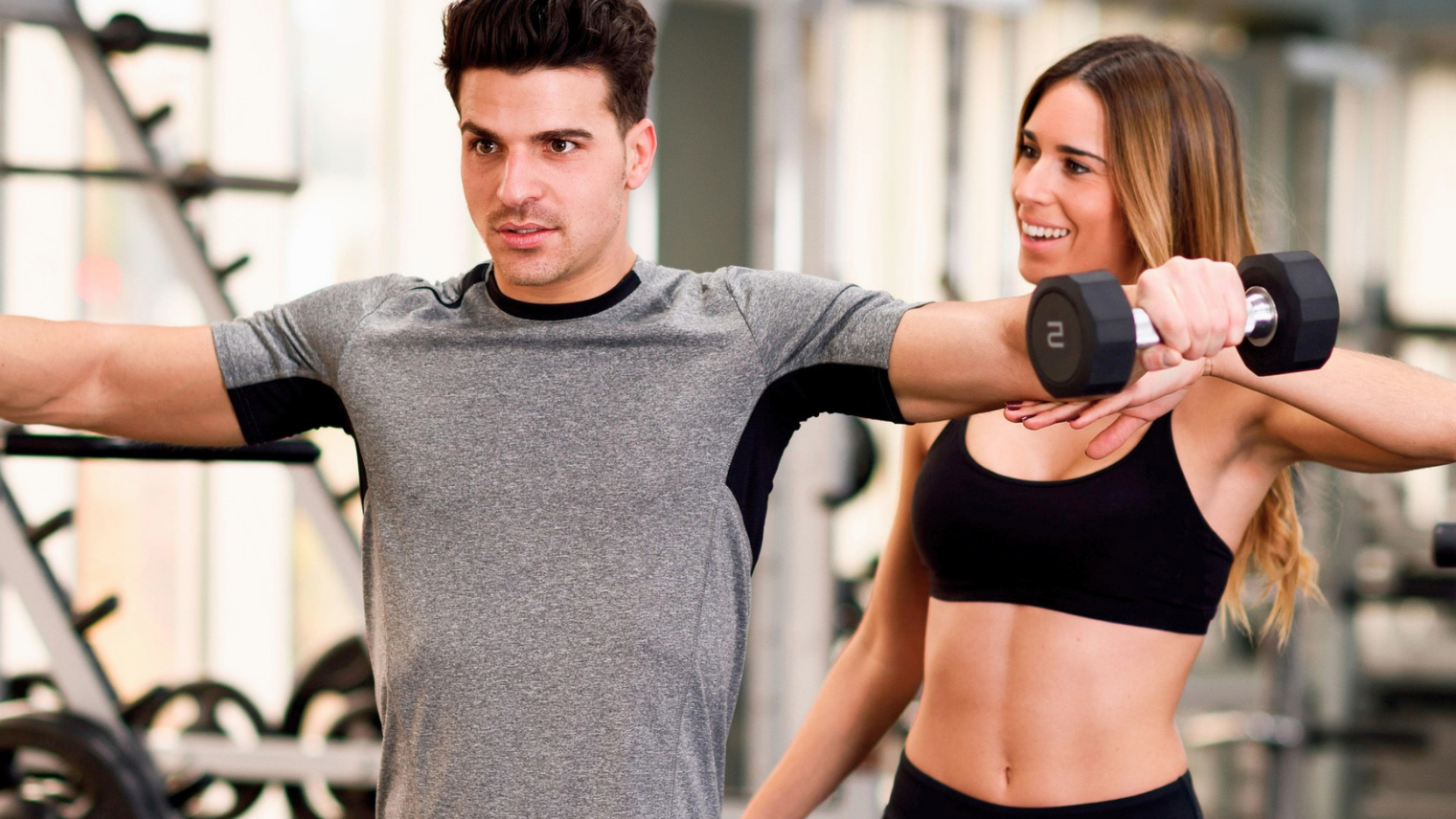 A man lifts his arms while a woman beside him holds a small dumbbell, both smiling in a gym.
