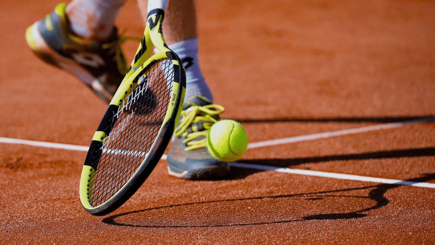 A tennis racket with a yellow and black frame hovers near a bouncing yellow ball on a reddish clay court, with a player’s shoes and white boundary lines visible.