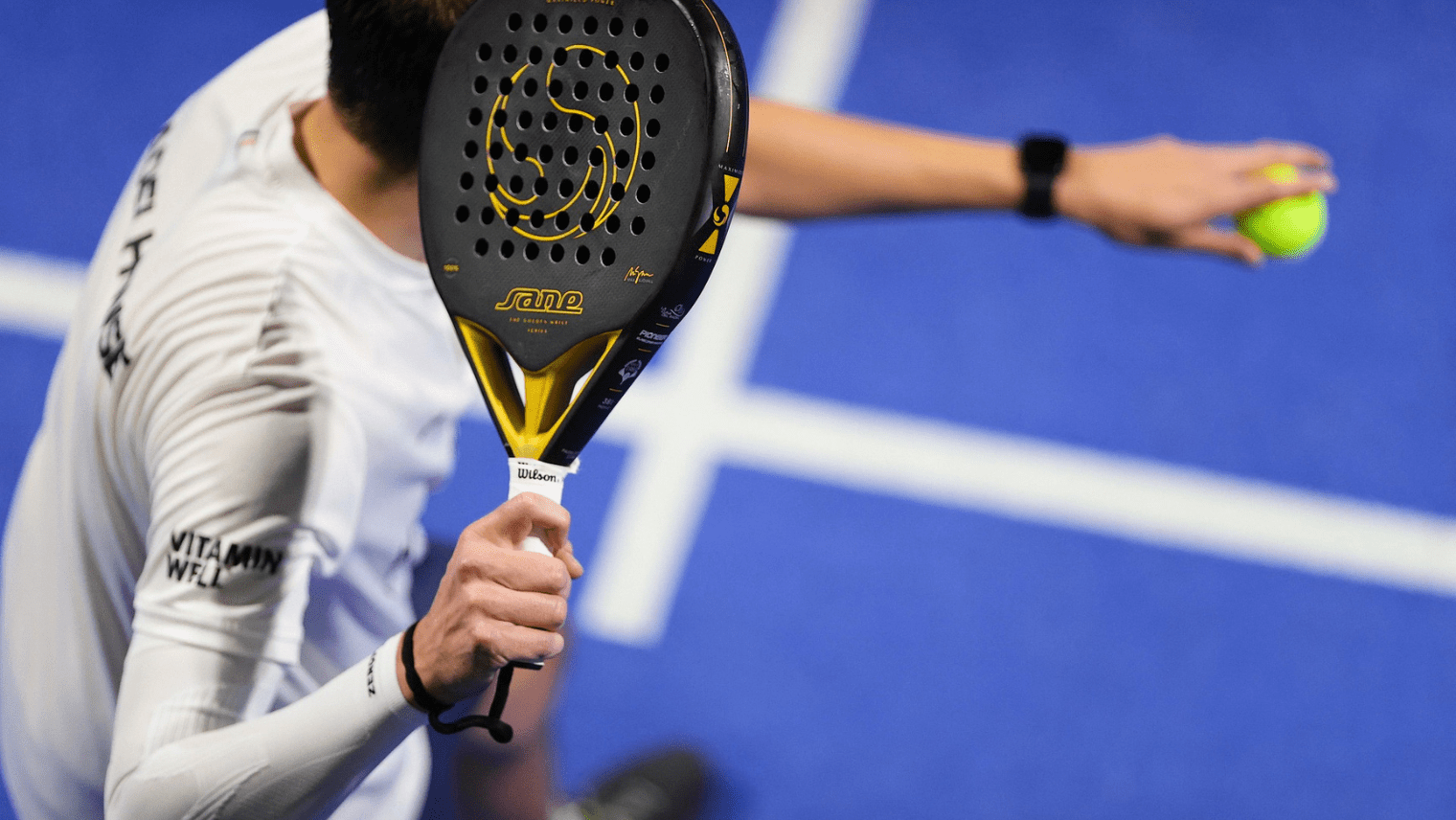 A person on a blue padel court holds a black and yellow perforated racket branded “SANE” and “Wilson,” ready to serve with a bright yellow ball in hand.