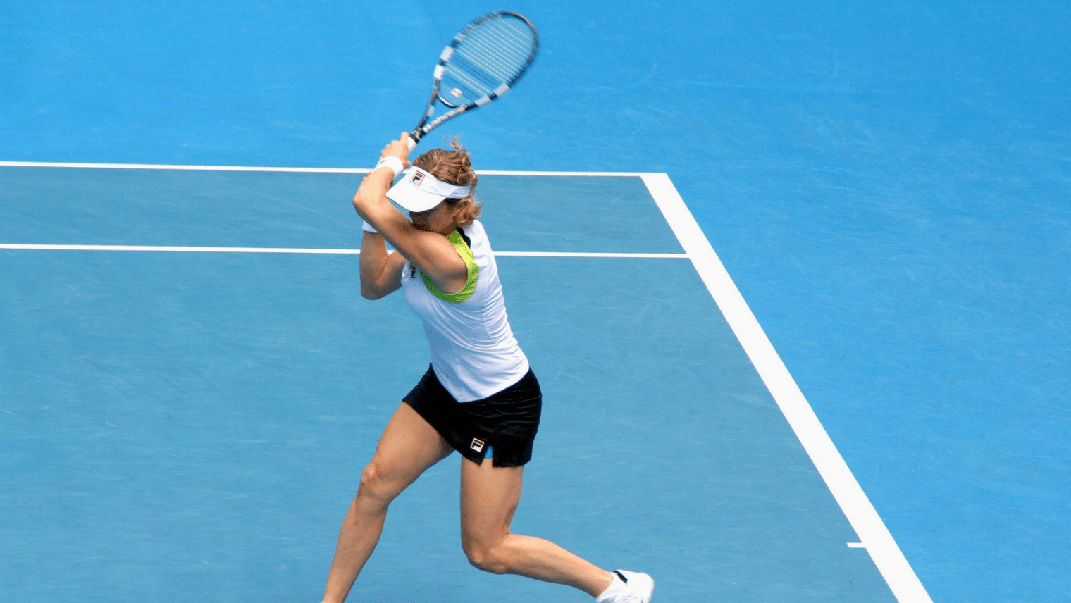 A female tennis player in a white sleeveless top and black shorts swings a backhand on a blue hard court, wearing a white visor and FILA-branded gear.