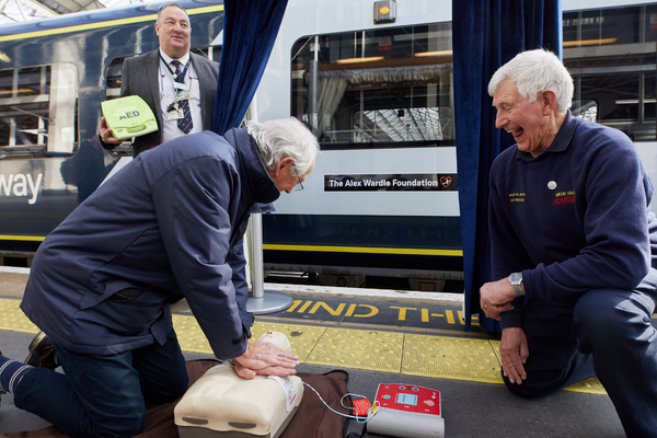 Two men kneeling on a platform beside a train with a resuscitation dummy between them