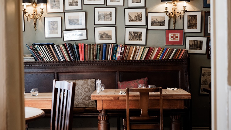 Body Image - The Wykeham Arms public house, interior. A row of books sits on a mantel, with chairs and tables in the foreground. The wall is covered with a number of hanging photos and images.